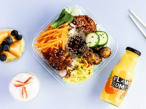 A bright salad box filled with rice, sweet potato and vegetables, accompanied by an orange juice drink, cheesecake pot, and fruit salad